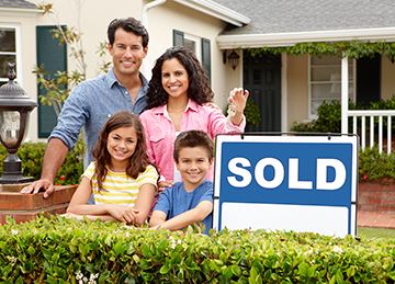 Two parents and two children pose next to a sold sign in front of a house