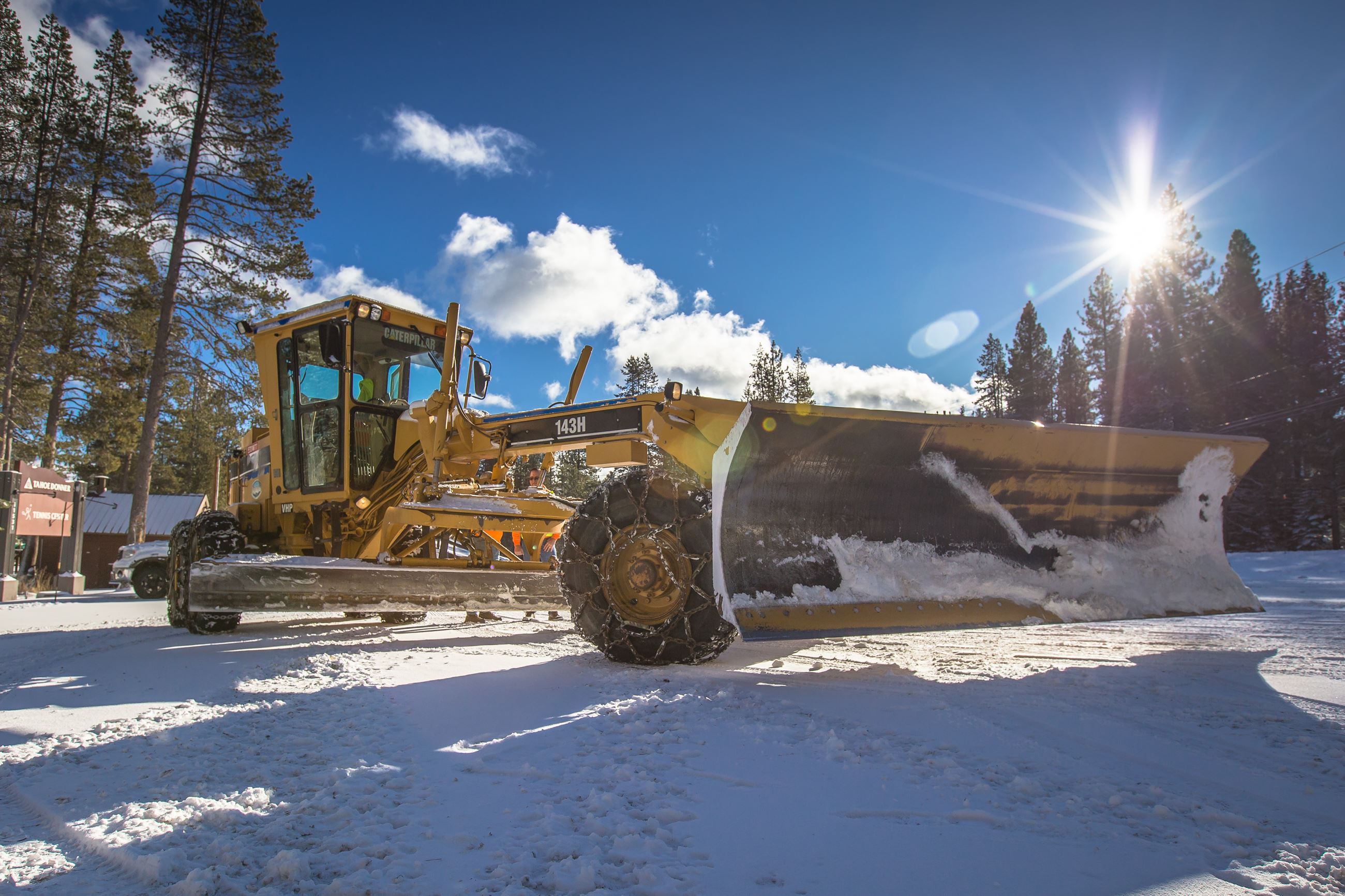 A large yellow snow grader driving in the snow with sun bursting through trees. 