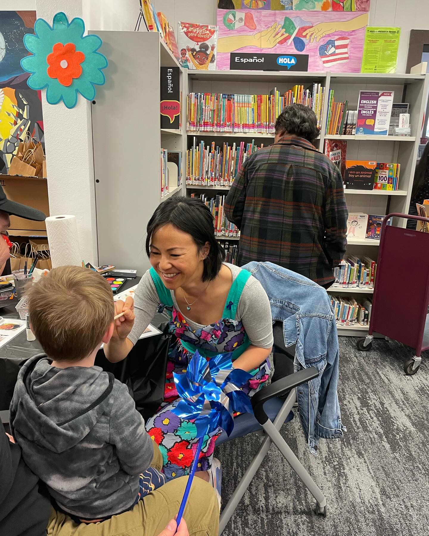 Women smiling as she paints a child's face in a library with people looking at books behind her. 