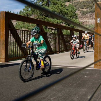Kids riding their bikes on a bridge with adults behind them on a sunny day. 